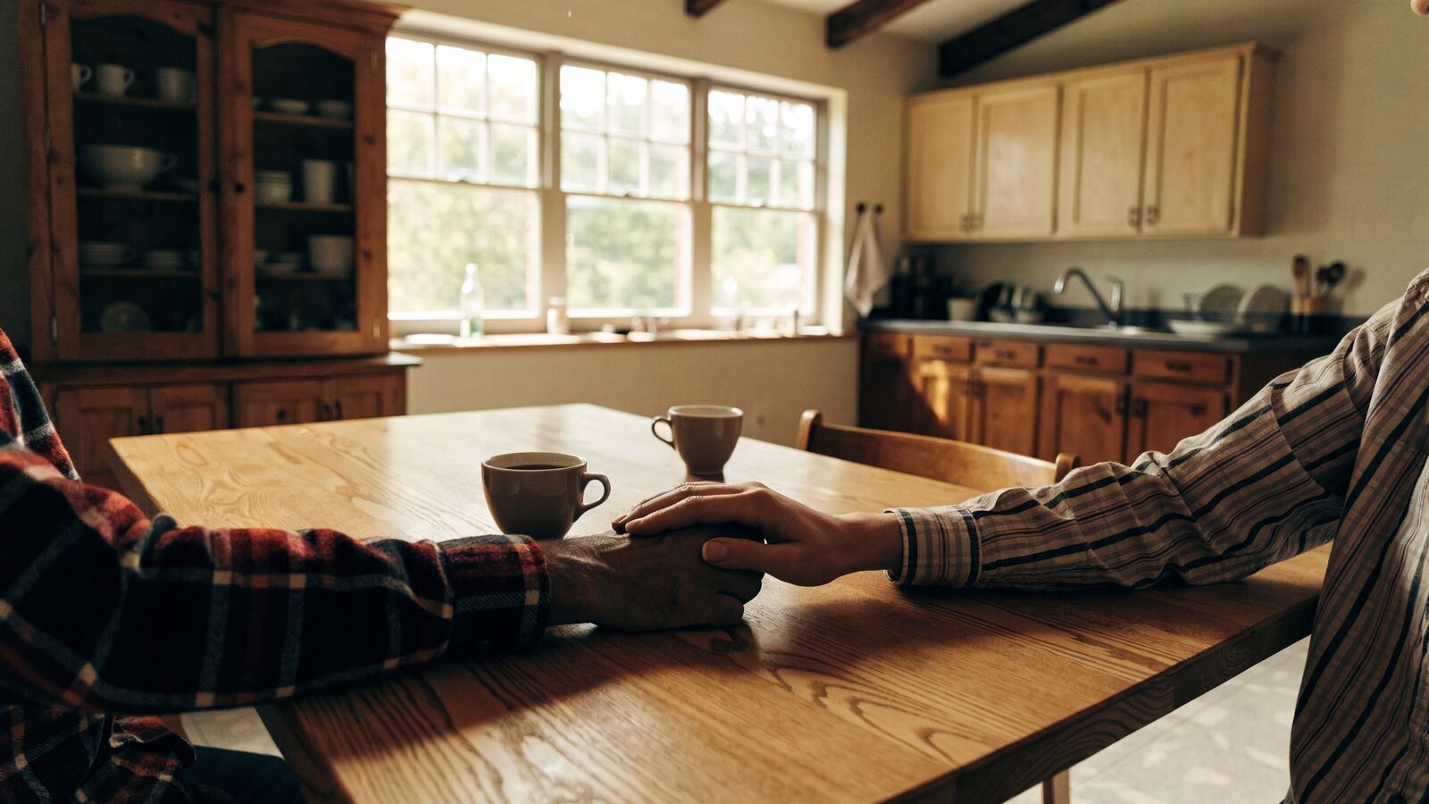 An adult child's hand resting reassuringly on an older parent's hand at a kitchen table