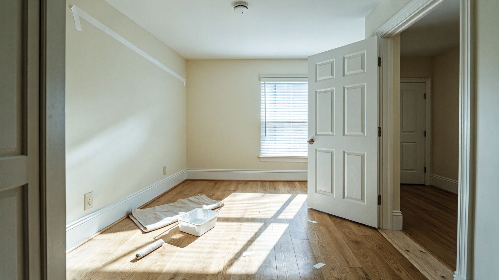 Painter's tape, a paint roller, and a drop cloth in a bright empty bedroom